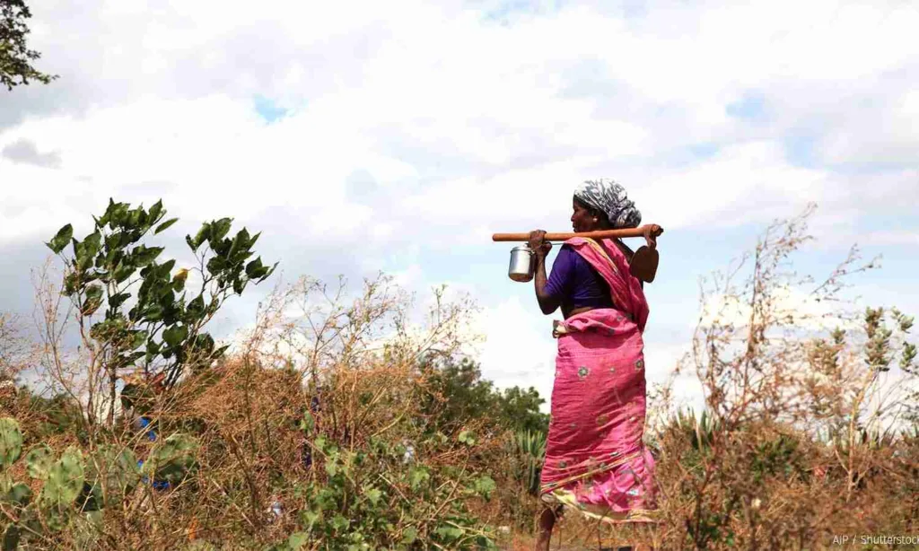 Two Indian working women in traditional sarees walking and working on a rural, unpaved path surrounded by vegetation, symbolizing the connection of rural women to land and agriculture.